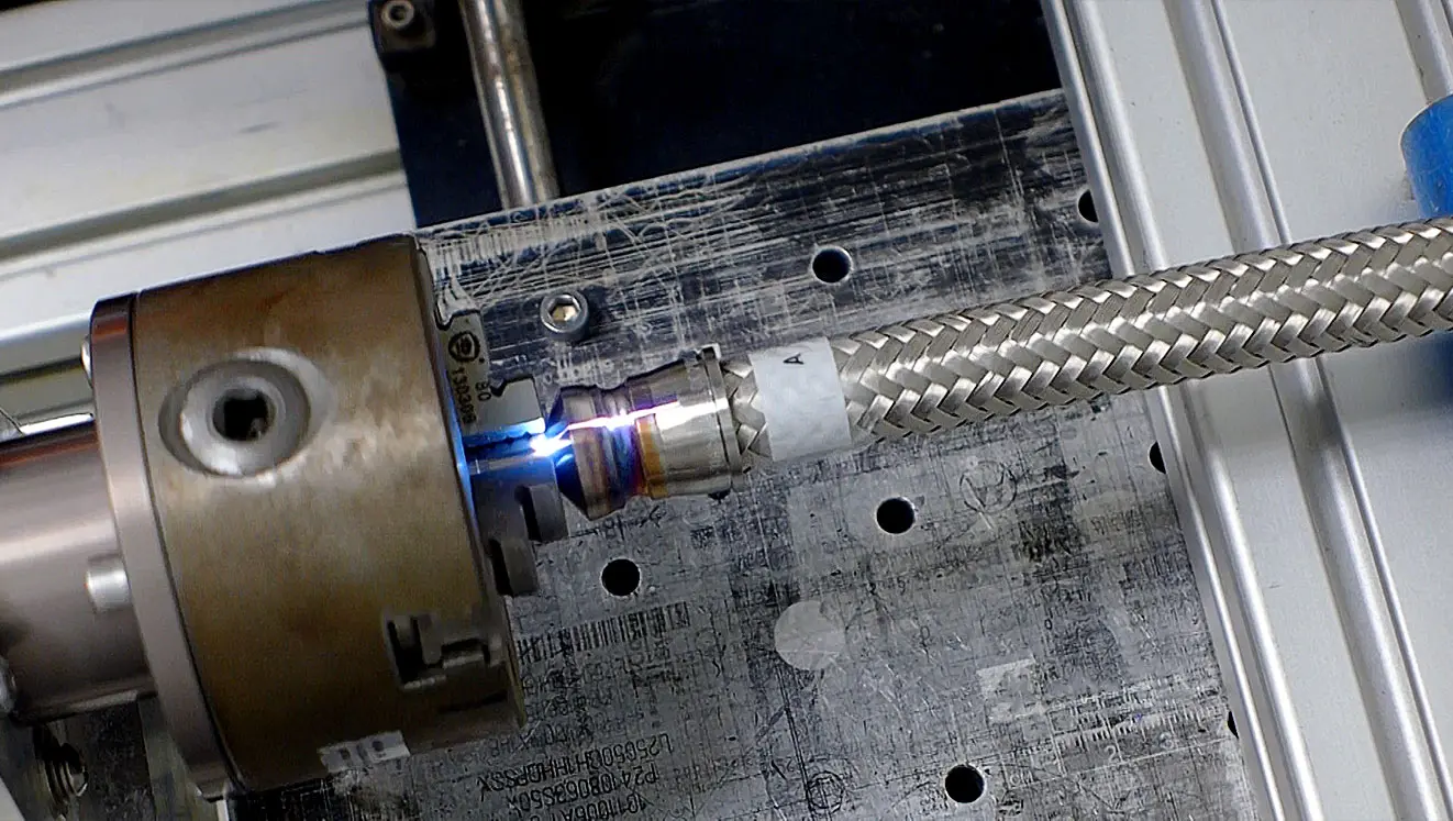 Close-up of a CMS Laser machine performing precision laser welding on a braided metal hose, with a bright blue laser beam joining the components, showcasing advanced industrial welding technology.